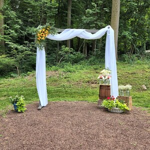 May include: A white fabric draped over a wooden archway decorated with sunflowers and greenery. The archway is set up outdoors in a grassy area with a wooden barrel and other floral arrangements.