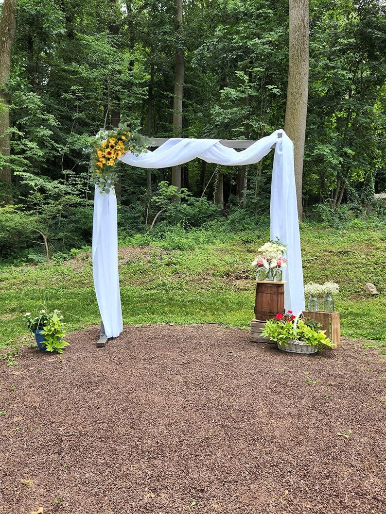 May include: A white fabric draped over a wooden archway decorated with sunflowers and greenery. The archway is set up outdoors in a grassy area with a wooden barrel and other floral arrangements.