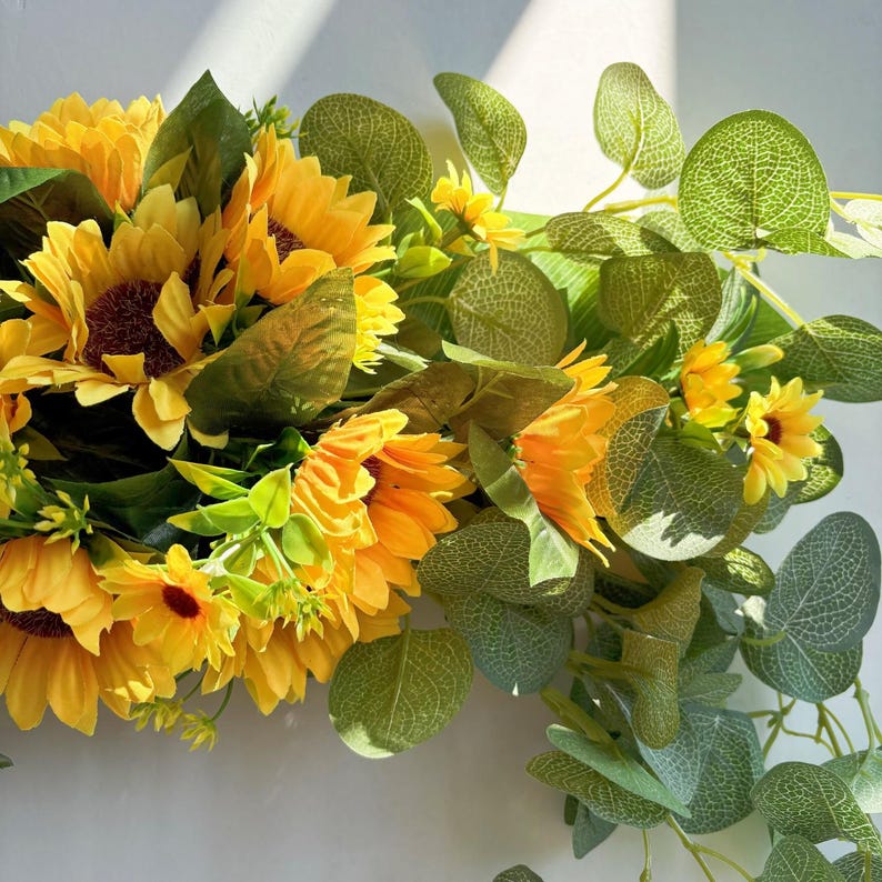 May include: A wreath made of artificial sunflowers and eucalyptus leaves. The sunflowers are yellow with brown centers and the eucalyptus leaves are green.