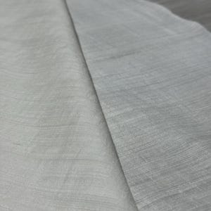 Puede incluir: Primer plano de dos secciones de tela, una gris claro y otra blanquecina. La tela tiene una apariencia sutilmente texturizada con l&iacute;neas horizontales visibles, lo que sugiere un material tejido. La iluminaci&oacute;n crea sombras suaves.