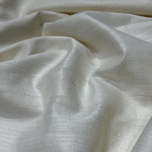 Puede incluir: Primer plano de tela blanquecina con un brillo sutil. El material est&aacute; drapeado, mostrando pliegues suaves y una superficie texturizada. La tela parece ligera y adecuada para ropa o decoraci&oacute;n del hogar.