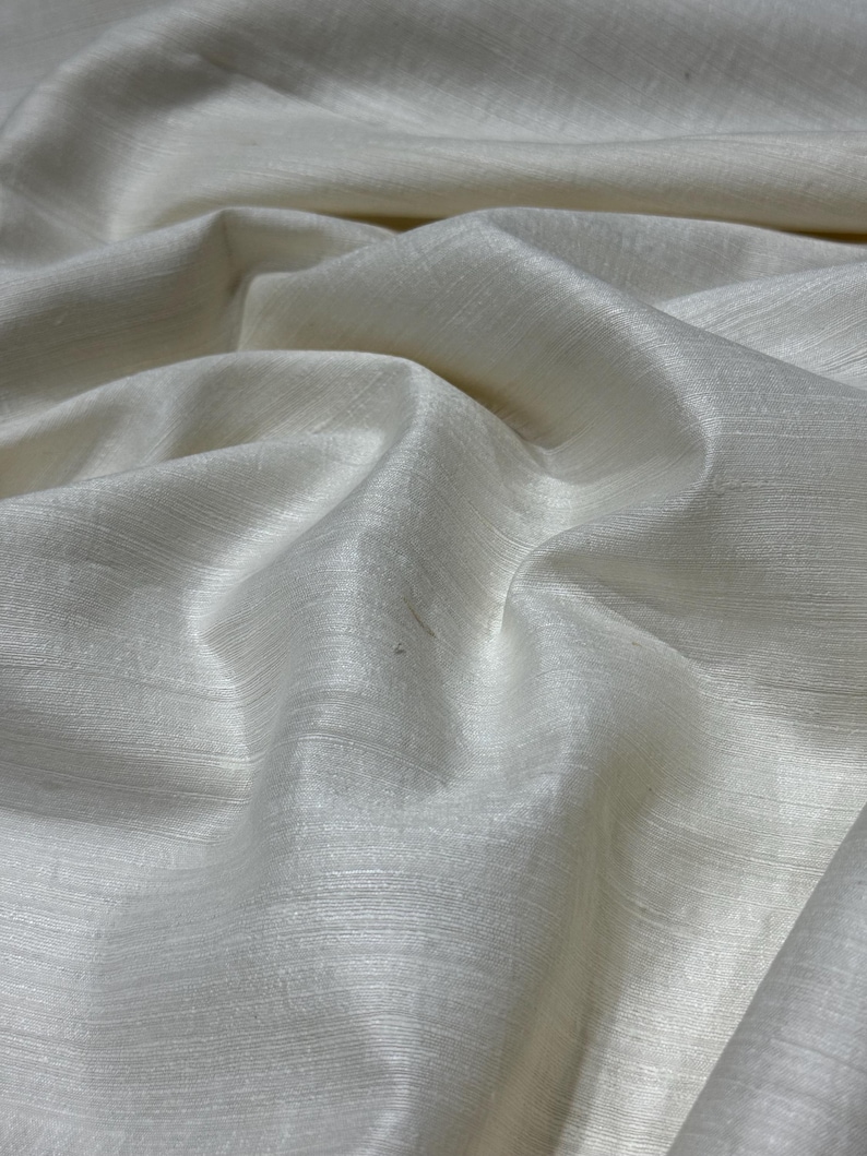 Puede incluir: Primer plano de tela blanquecina con un brillo sutil. El material est&aacute; drapeado, mostrando pliegues suaves y una superficie texturizada. La tela parece ligera y adecuada para ropa o decoraci&oacute;n del hogar.