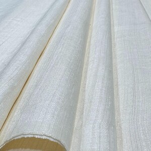 Puede incluir: Primer plano de tela blanquecina con tejido texturizado. La tela est&aacute; drapeada en pliegues suaves, creando sombras y reflejos. El material parece ligero y adecuado para ropa o decoraci&oacute;n del hogar.