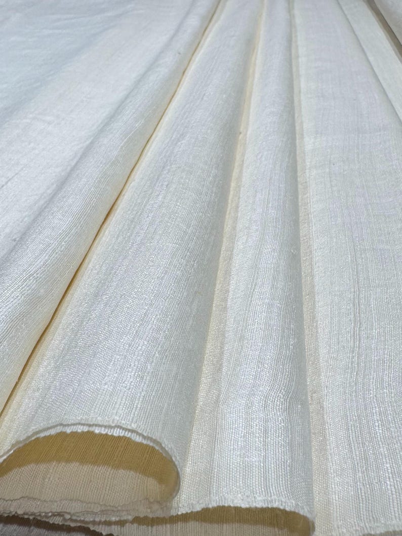 Puede incluir: Primer plano de tela blanquecina con tejido texturizado. La tela est&aacute; drapeada en pliegues suaves, creando sombras y reflejos. El material parece ligero y adecuado para ropa o decoraci&oacute;n del hogar.