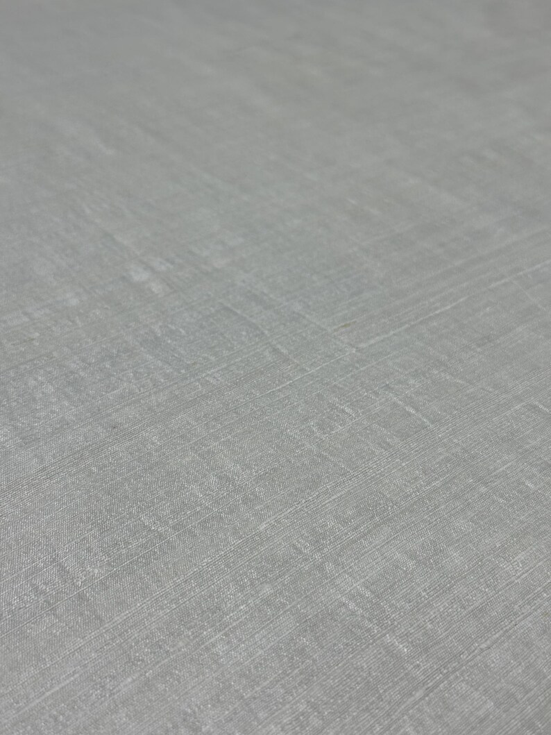 Puede incluir: Primer plano de una tela gris claro con un tejido sutil y texturizado. La superficie de la tela tiene una calidad ligeramente iridiscente, con l&iacute;neas diagonales y horizontales visibles. La apariencia general es suave y refinada.