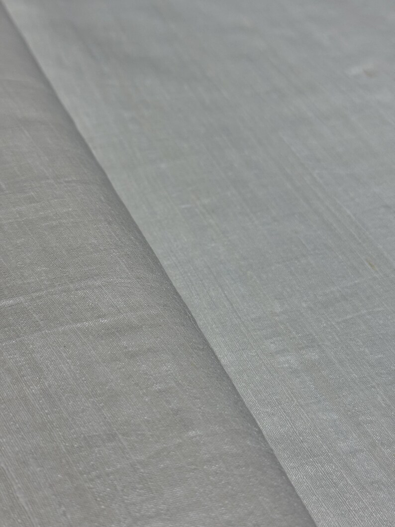 Puede incluir: Primer plano de una tela gris claro con un tejido sutil y texturizado. La tela est&aacute; doblada, creando l&iacute;neas diagonales y sombras. El material parece suave y ligero, adecuado para ropa o decoraci&oacute;n del hogar.