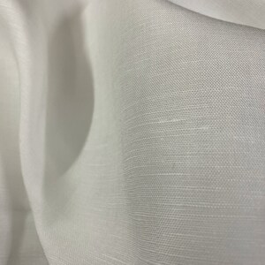 May include: White linen fabric with a smooth, woven texture. The fabric is folded and draped, showing its soft, flowing quality.