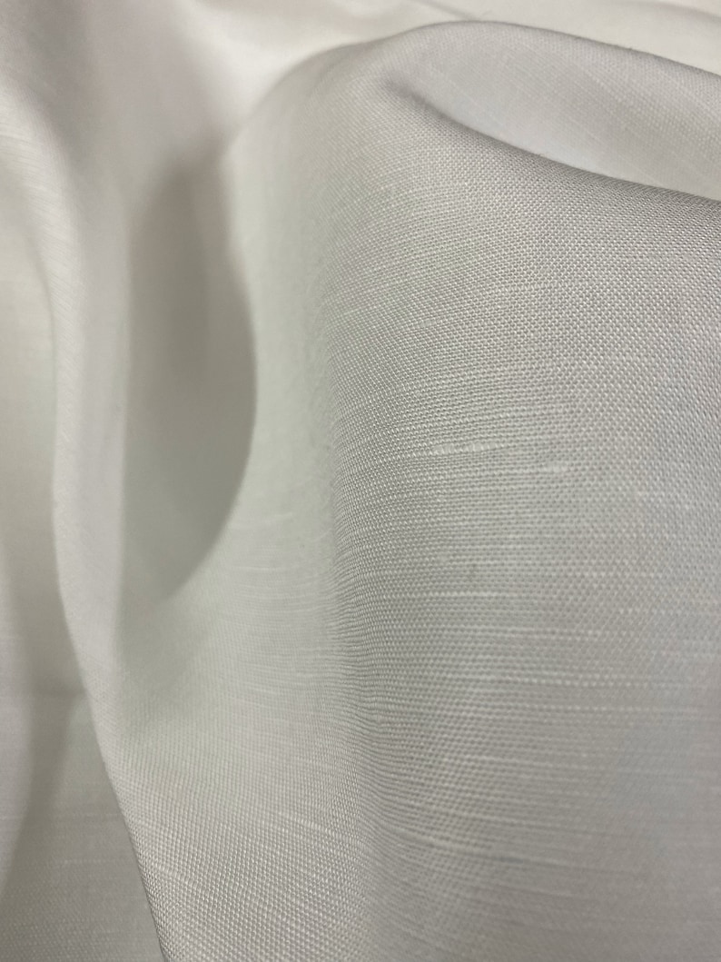 May include: White linen fabric with a smooth, woven texture. The fabric is folded and draped, showing its soft, flowing quality.
