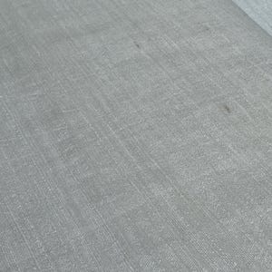 Puede incluir: Primer plano de una tela gris claro con un tejido texturizado. La tela tiene un brillo sutil y parece ser lino o un material similar. La superficie muestra ligeras variaciones de color y textura.