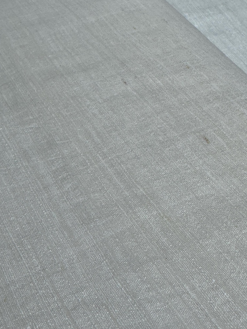 Puede incluir: Primer plano de una tela gris claro con un tejido texturizado. La tela tiene un brillo sutil y parece ser lino o un material similar. La superficie muestra ligeras variaciones de color y textura.