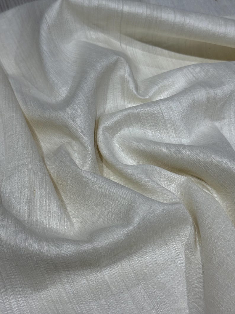 Puede incluir: Primer plano de una tela color crema con un brillo sutil. El material est&aacute; drapeado en pliegues suaves, creando una superficie texturizada. La tela parece ligera y fluida, con l&iacute;neas verticales visibles.