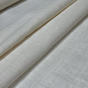 Puede incluir: Primer plano de tela blanquecina con un tejido sutil. La tela est&aacute; doblada, creando sombras suaves y resaltando la ca&iacute;da del material. El color es un tono cremoso y neutro, adecuado para diversos proyectos textiles.