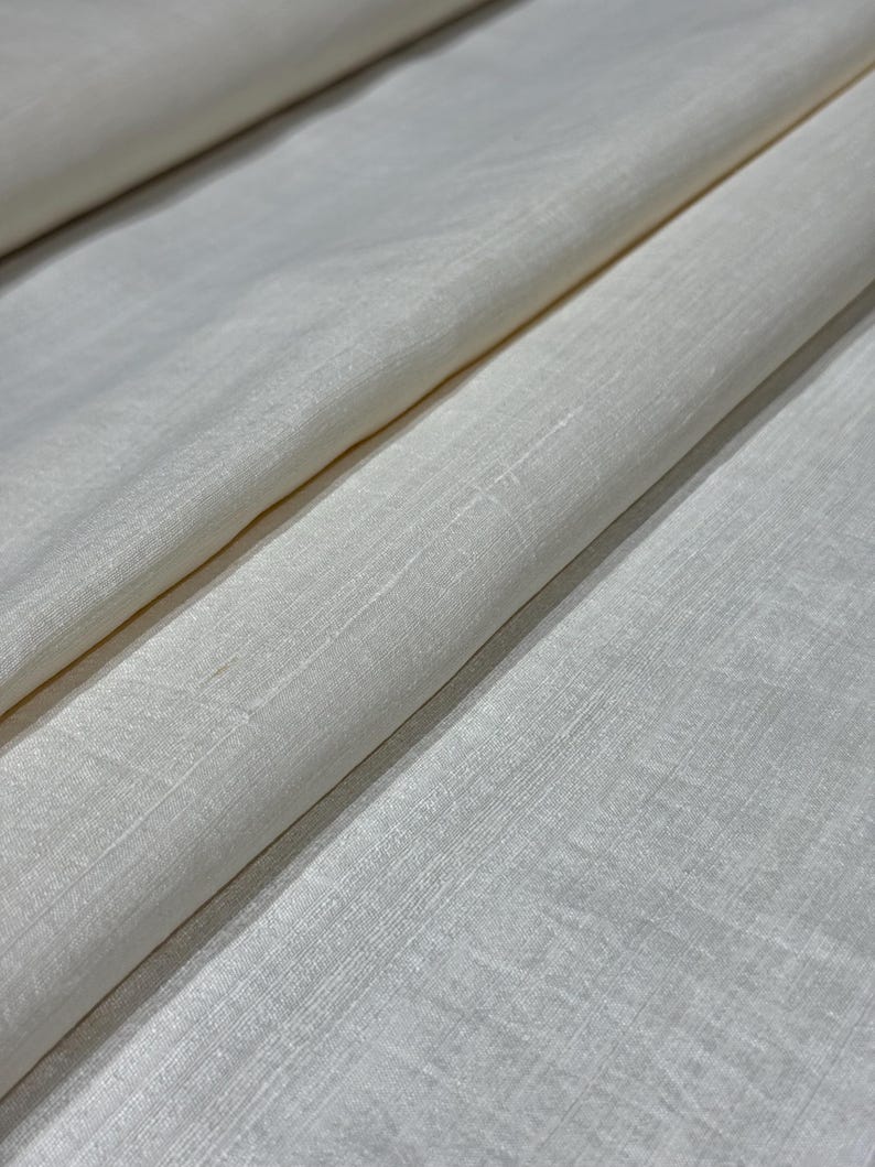 Puede incluir: Primer plano de tela blanquecina con un tejido sutil. La tela est&aacute; doblada, creando sombras suaves y resaltando la ca&iacute;da del material. El color es un tono cremoso y neutro, adecuado para diversos proyectos textiles.