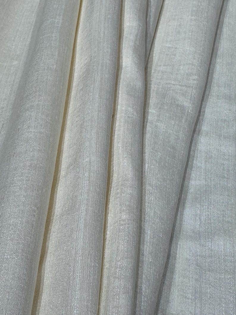 Puede incluir: Primer plano de tela blanquecina con l&iacute;neas verticales y textura sutil. La tela est&aacute; drapeada, creando pliegues y sombras suaves. El material parece ser ligero, posiblemente lino o algod&oacute;n, adecuado para ropa o decoraci&oacute;n del hogar.