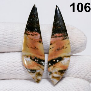 May include: Two polished gemstone pieces with a leaf-like shape. The stones feature a mix of colors, including black, peach, yellow, and white. The number "106" is visible in the upper right corner. The stones are displayed on a white surface.