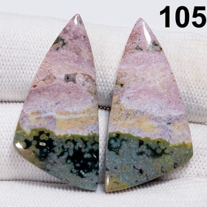 May include: Two polished, triangular gemstones with a layered appearance. The stones feature a blend of colors, including pink, white, yellow, green, and black. The number "105" is visible in the upper right corner. These stones could be used for jewelry making.