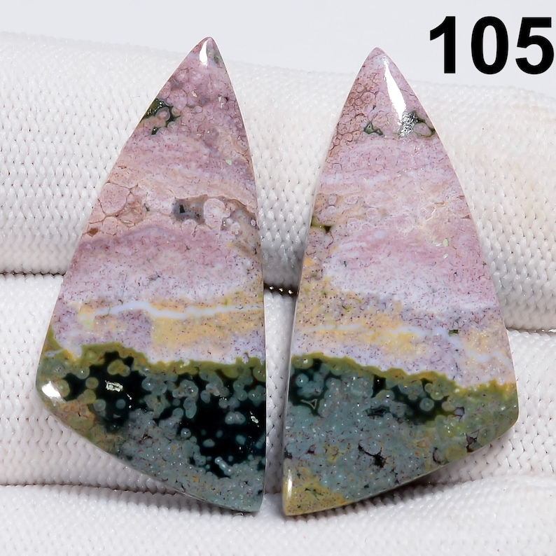 May include: Two polished, triangular gemstones with a layered appearance. The stones feature a blend of colors, including pink, white, yellow, green, and black. The number "105" is visible in the upper right corner. These stones could be used for jewelry making.