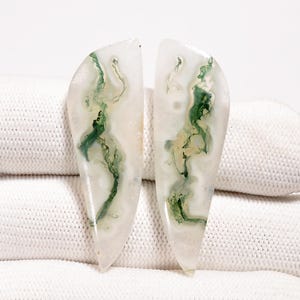 Può includere: Due pietre preziose lucide a forma di goccia. Le pietre sono di colore bianco traslucido con intricate inclusioni verdi simili a muschio. Le pietre preziose sono esposte su una superficie bianca strutturata.