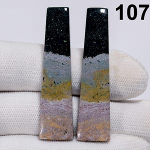 May include: Two rectangular gemstones with a layered appearance. The top section is black with white speckles, followed by gray, yellow, and lavender bands. The number "107" is visible in the upper right corner.
