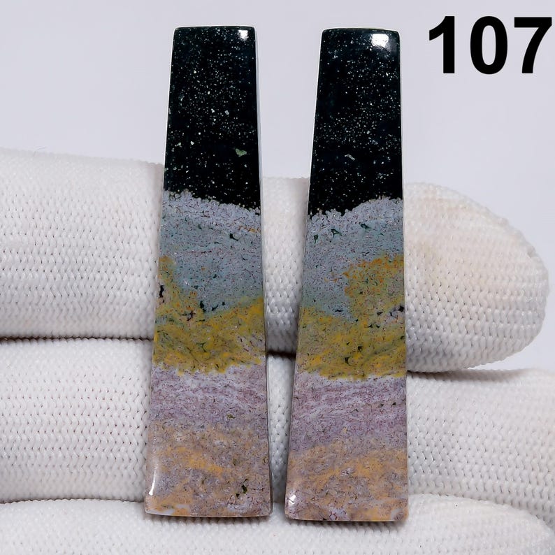 May include: Two rectangular gemstones with a layered appearance. The top section is black with white speckles, followed by gray, yellow, and lavender bands. The number "107" is visible in the upper right corner.