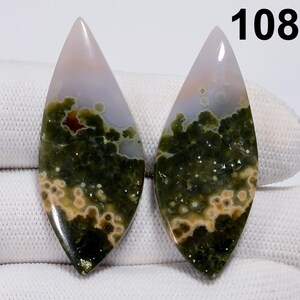 May include: Two polished gemstone cabochons with a teardrop shape. The stones feature a mix of green, beige, and white patterns. The top of each stone is a pale pink color. The number "108" is visible in the upper right corner.