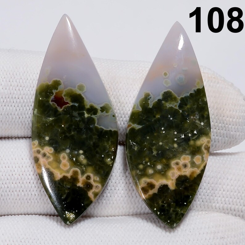 May include: Two polished gemstone cabochons with a teardrop shape. The stones feature a mix of green, beige, and white patterns. The top of each stone is a pale pink color. The number "108" is visible in the upper right corner.