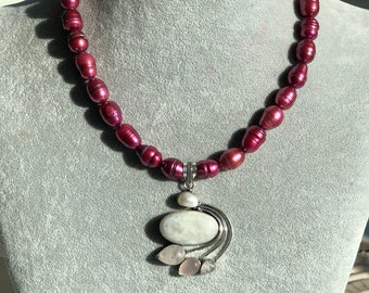 May include: A necklace made of deep burgundy pearls with a silver pendant featuring a large white oval stone and smaller pink stones.