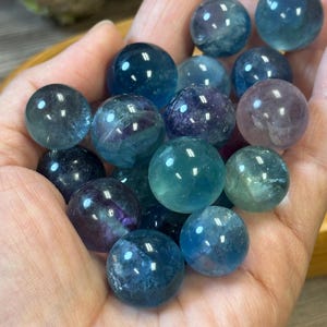 May include: A collection of polished fluorite spheres in shades of blue, green, and purple. The spheres are held in a hand, showcasing their translucent quality and varied colour patterns. The spheres are approximately 2.5 cm in diameter.