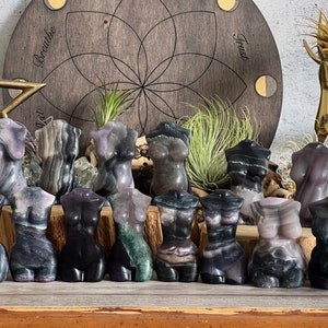Op de afbeelding: Een verzameling van 12 kleine, gebeeldhouwde sculpturen van vrouwelijke torso's gemaakt van verschillende soorten gebande agaat. De sculpturen zijn in tinten paars, zwart en groen.