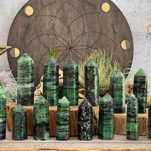 Könnte beinhalten: Eine Sammlung von 15 grünen und schwarz gestreiften Edelsteintürmen, auch bekannt als grüner Aventurin, angeordnet auf einer Holzoberfläche. Die Türme sind alle unterschiedlich groß und geformt, und einige haben einen ausgeprägteren schwarzen Streifen als andere.