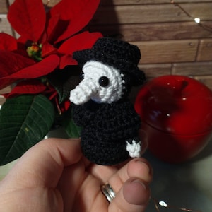 May include: A small crocheted black and white figure wearing a black hat. The figure has a long white nose and is holding a small white object in its hand. The figure is being held in a hand with a ring on the finger.