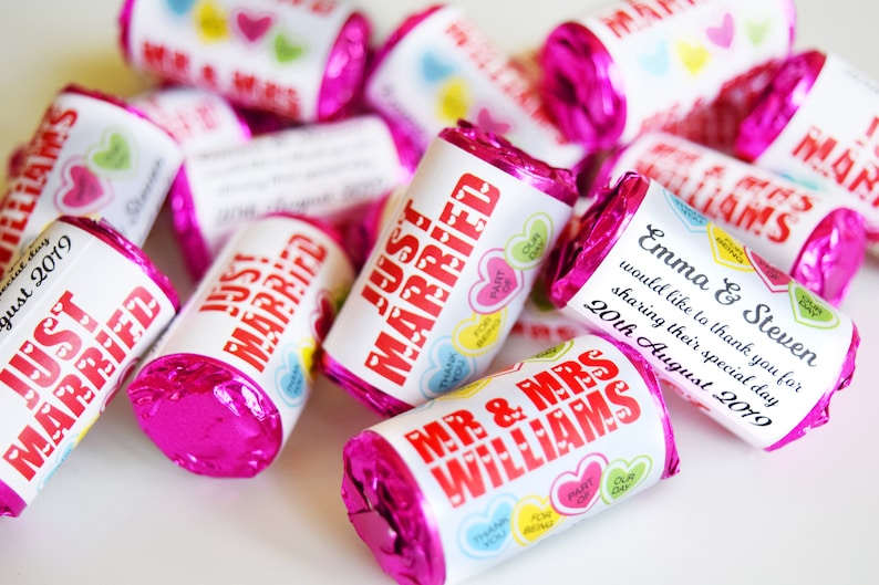 May include: A collection of cylindrical chocolate candies wrapped in bright pink foil. Each candy features a white paper label printed with red text reading Just Married or Mr & Mrs Williams, alongside small colorful heart graphics.