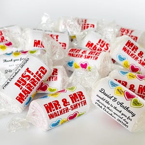 May include: A collection of individually wrapped candies featuring white labels with red text reading "JUST MARRIED" or "MR & MR WALKER-SMYTH" and "David & Anthony". The labels are decorated with small colorful heart graphics and wedding dates.