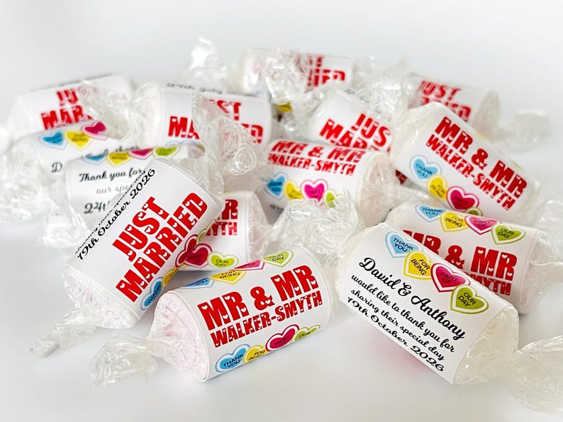 May include: A collection of individually wrapped candies featuring white labels with red text reading "JUST MARRIED" or "MR & MR WALKER-SMYTH" and "David & Anthony". The labels are decorated with small colorful heart graphics and wedding dates.