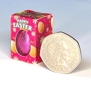 May include: A small pink chocolate egg in a pink and white box with the words "Happy Easter" printed on it. The box is next to a British 50 pence coin for size comparison.