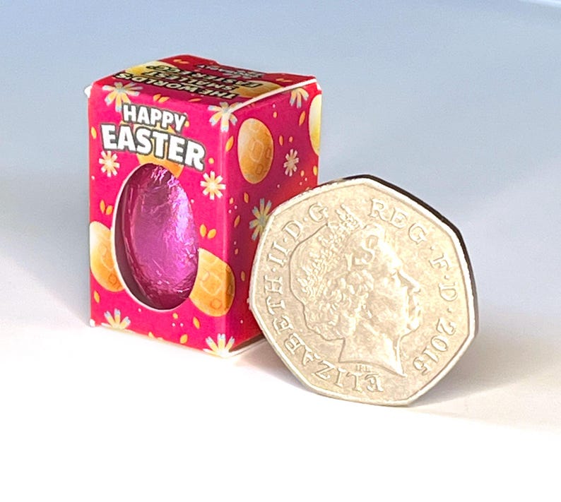 May include: A small pink chocolate egg in a pink and white box with the words "Happy Easter" printed on it. The box is next to a British 50 pence coin for size comparison.