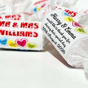 May include: Cylindrical candy rolls wrapped in clear plastic with personalized white labels. One label reads MR & MRS WILLIAMS with heart graphics. Another label reads Harry & Emma would like to thank you for sharing their special day 30th August 2026.