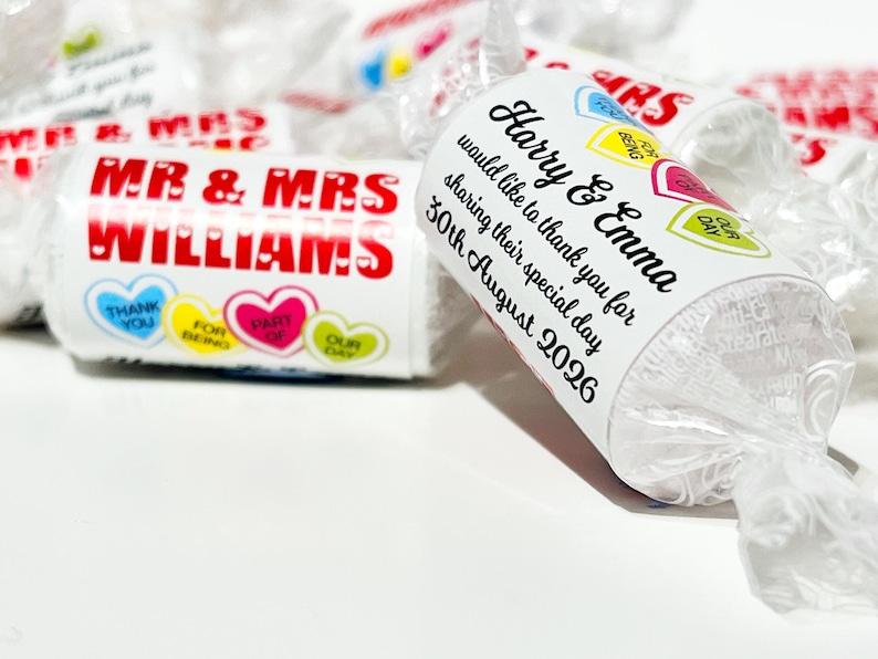May include: Cylindrical candy rolls wrapped in clear plastic with personalized white labels. One label reads MR & MRS WILLIAMS with heart graphics. Another label reads Harry & Emma would like to thank you for sharing their special day 30th August 2026.
