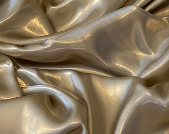 May include: Close-up of a shimmering, draped fabric in a light champagne color. The material has a glossy sheen, with soft folds and highlights that create a luxurious appearance. Ideal for fashion or home decor.