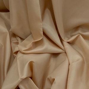 May include: Close-up of beige fabric with soft folds and a silky sheen. The fabric is draped, creating shadows and highlights that emphasise its texture and flow. The colour is a warm, neutral tone.