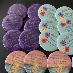 May include: Assortment of colorful round buttons with inspirational quotes and floral designs. The quotes include "Rejoice in the Lord always", "Above all love each other", and "For by grace you have been saved through faith".