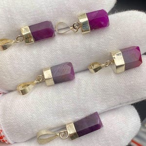 May include: Five faceted, rectangular pendants with a deep magenta hue. Each pendant has a silver-colored metal cap and a loop for a chain. The pendants are displayed on a white surface.