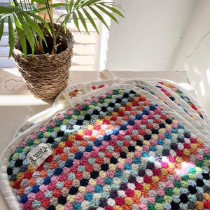 May include: A stack of colourful, handmade crochet squares with a white border. The squares feature a mix of bright colours, including pink, blue, green, and orange. A small tag with the text "DAMLA ROSE" is attached. A potted plant is in the background.