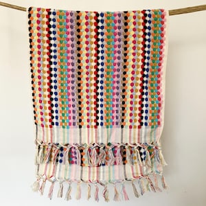 Könnte beinhalten: Ein farbenfrohes, gemustertes Handtuch mit Fransen. Das Handtuch zeigt vertikale Reihen mehrfarbiger Punkte in Rot-, Blau-, Gelb-, Grün- und Lilatönen. Der untere Rand hat ein Streifenmuster und eine Fransen aus geknoteten Quasten.