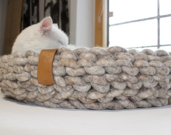 May include: A white cat sleeps in a large, round, gray and white knitted pet bed with a brown leather tag.