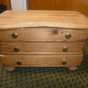 Könnte beinhalten: Eine dreischübige Holzkiste mit abgerundeter Oberseite und Messingknöpfen. Die Kiste ist aus hellem braunem Holz gefertigt und hat eine Antik-Optik.