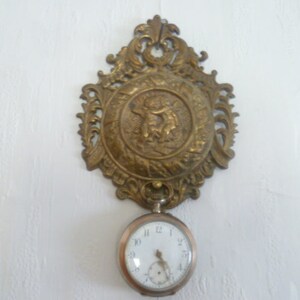 Puede incluir: Un reloj de pared antiguo de latón con un marco ornamentado. El marco tiene un medallón central con un relieve y una parte superior decorativa. Un reloj de bolsillo plateado cuelga debajo. El reloj está montado en una pared blanca.