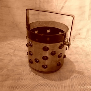 Puede incluir: Un cubo de hielo de plata con una parte superior redonda abierta y un asa. El cubo está decorado con pequeños acentos redondos de color marrón oscuro.