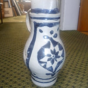 May include: A salt-glazed stoneware pitcher with a light gray body and cobalt blue hand-painted floral and geometric patterns. The vessel features a bulbous body, a cylindrical neck with a pouring spout, and a single side handle for gripping.