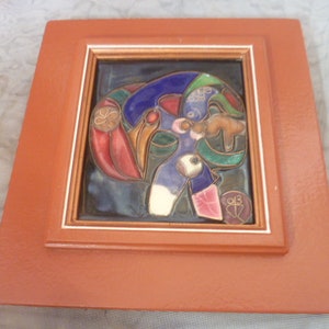 May include: A square wall plaque featuring a central ceramic tile with a colorful abstract design in blue, red, green, and white, encased in a thick, matte orange wooden frame with a thin gold-toned inner border and a small circular seal.